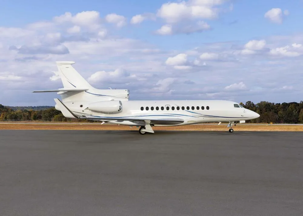 A white Dassault Falcon business jet with blue stripes seen from the side on an airfield.