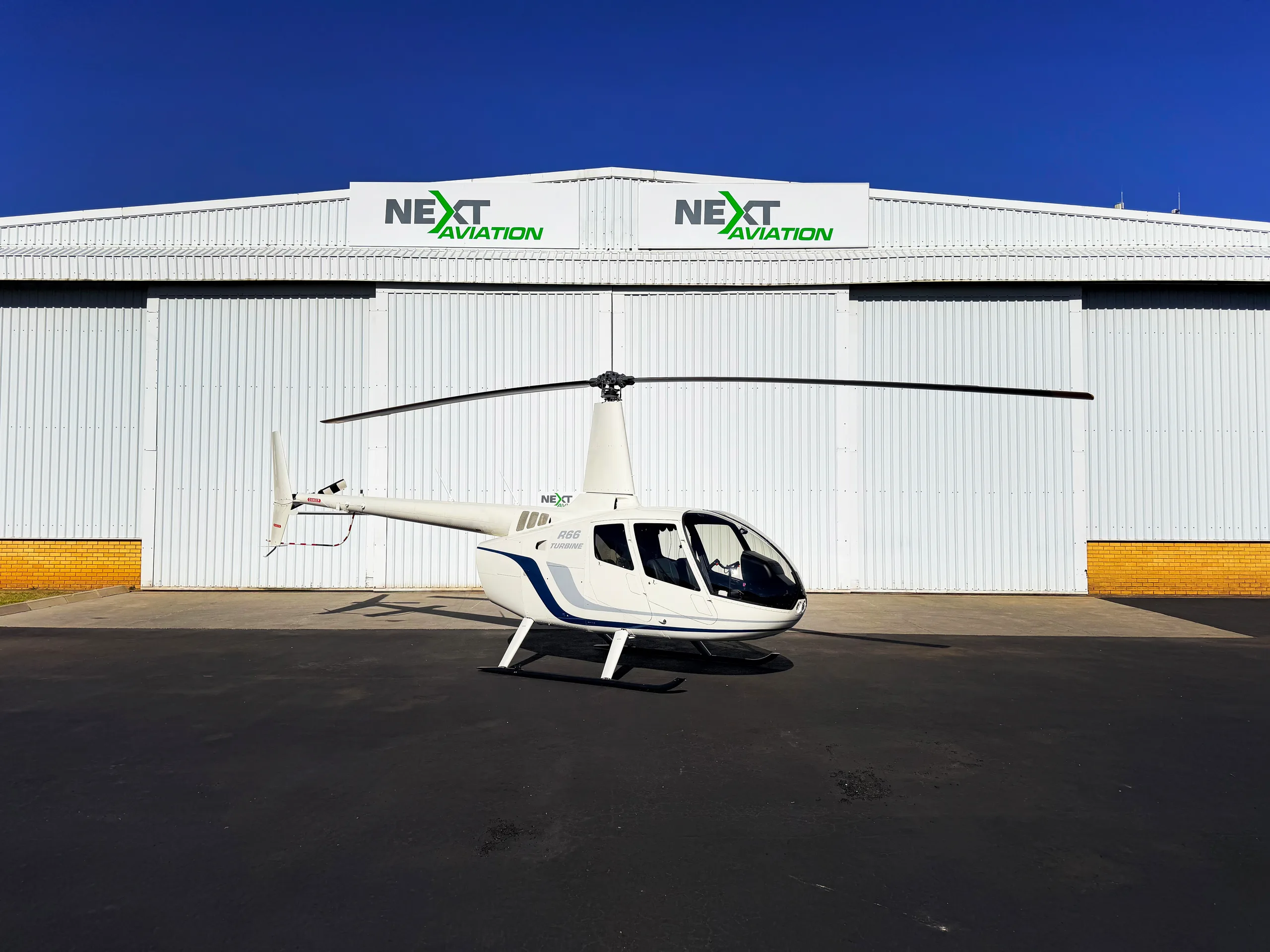 A white Robinson R66 Turbine helicopter with blue and grey stripes is parked on a tarmac in front of a white hangar with "NEXT AVIATION" logos.