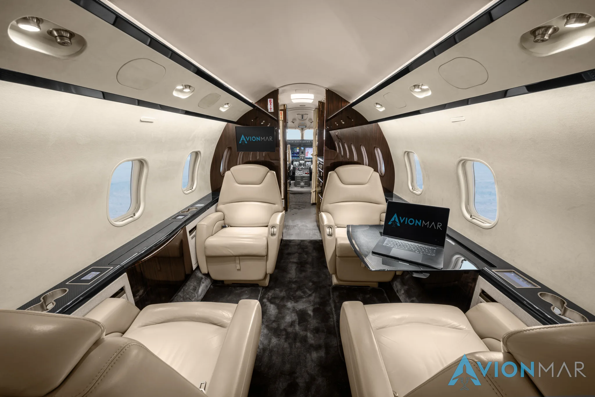 The luxurious interior of a private jet cabin with cream leather seats and a laptop on a table, viewed from the rear.