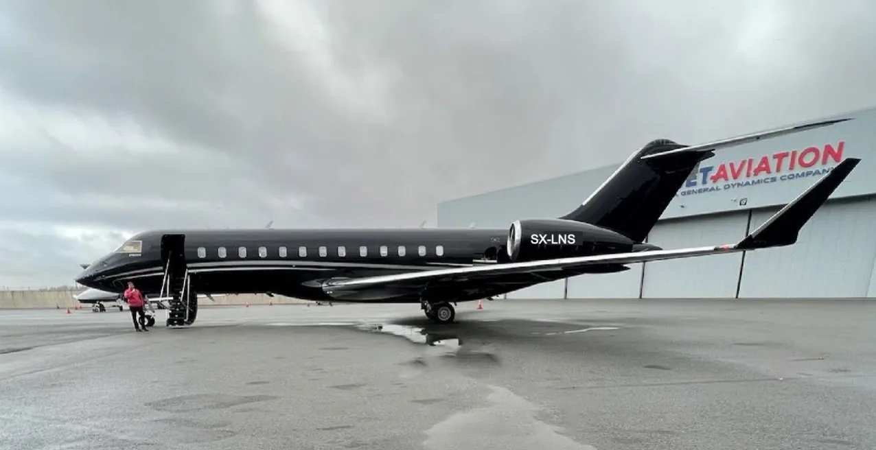 A black Bombardier Global Express XRS aircraft with registration SX-LNS parked on a wet tarmac in front of a hangar, with a person near the open door.