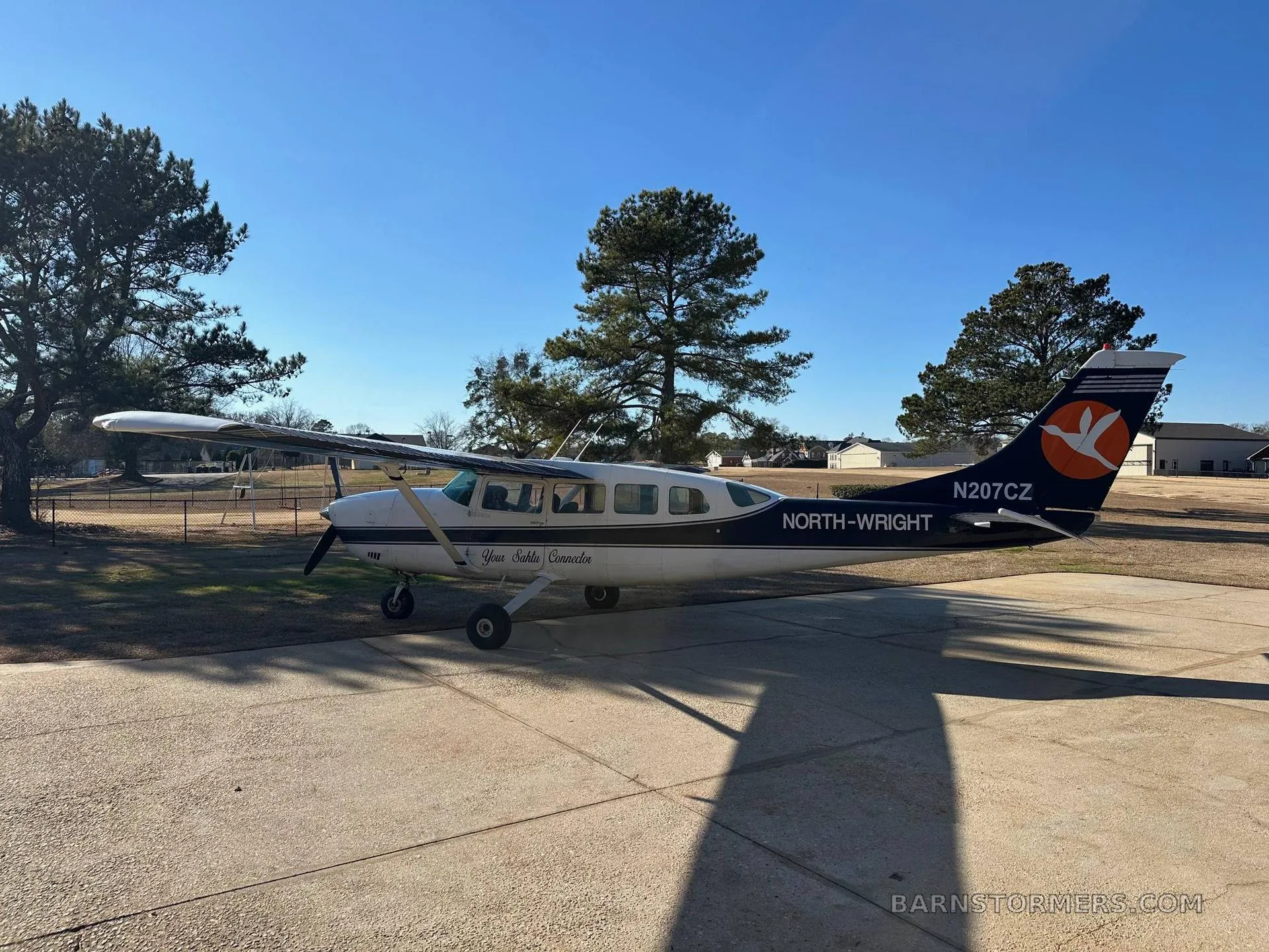 A white and dark blue Cessna 207 aircraft, N207CZ, with an orange bird logo on its tail, parked on a concrete driveway surrounded by trees.
