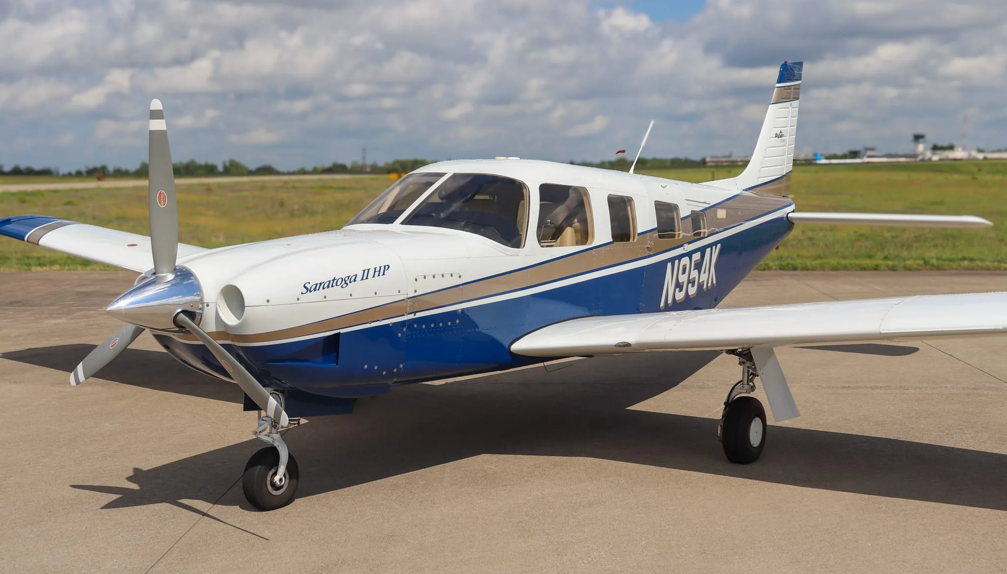 A Piper Saratoga II HP aircraft, N954K, in white, blue, and gold livery, parked on an airfield.
