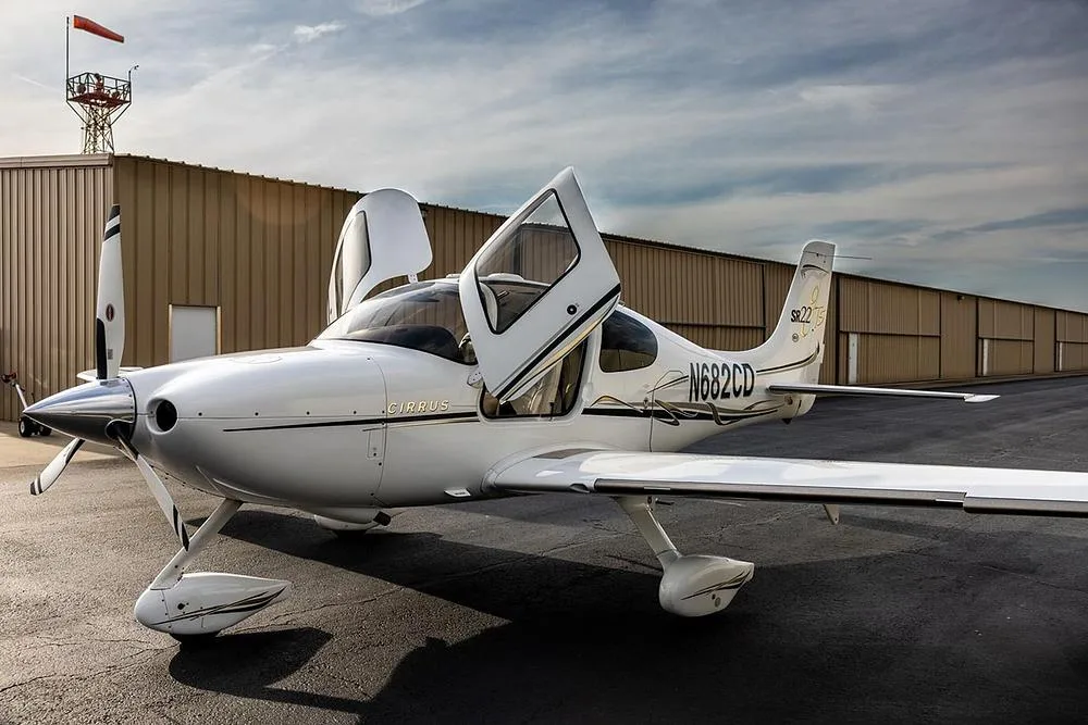 A white Cirrus SR22T aircraft with black and gold accents, registration N682CD, parked on an airfield with its doors open.