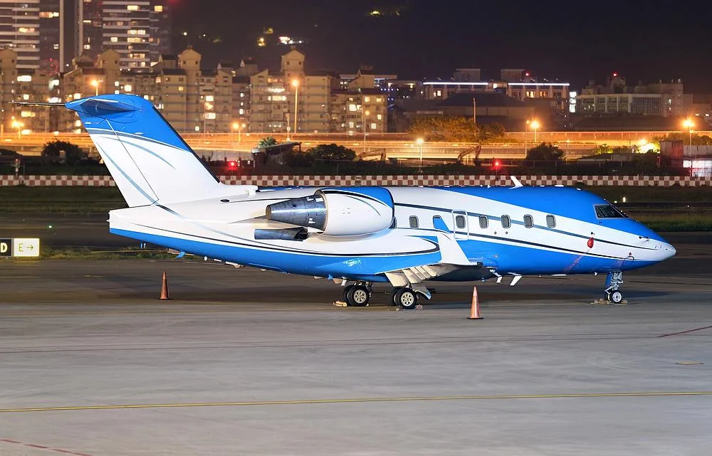 A blue and white Bombardier Challenger 604 private jet is parked on an airport tarmac at night, illuminated by city lights.