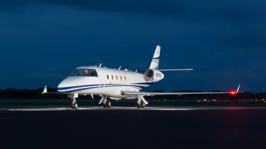 A white business jet with blue and grey stripes is parked on a tarmac at night, with its landing lights illuminated.