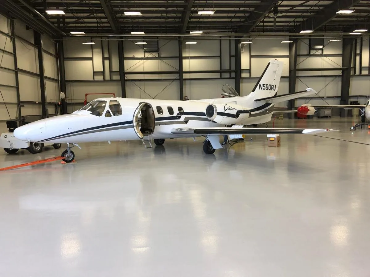 A white Cessna Citation business jet with black and grey stripes is parked inside a hangar with its main door open.