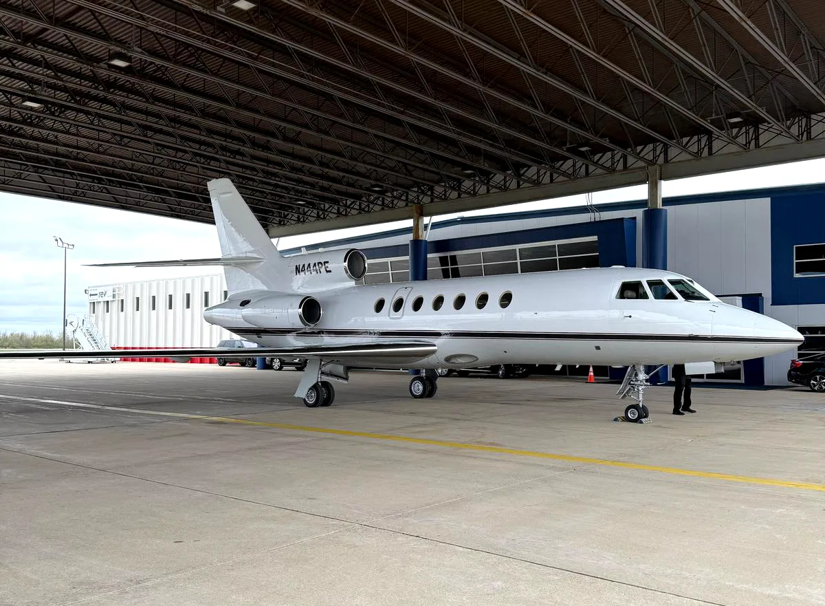 A white Dassault Falcon business jet, registration N444PE, parked under a hangar structure at an airport.
