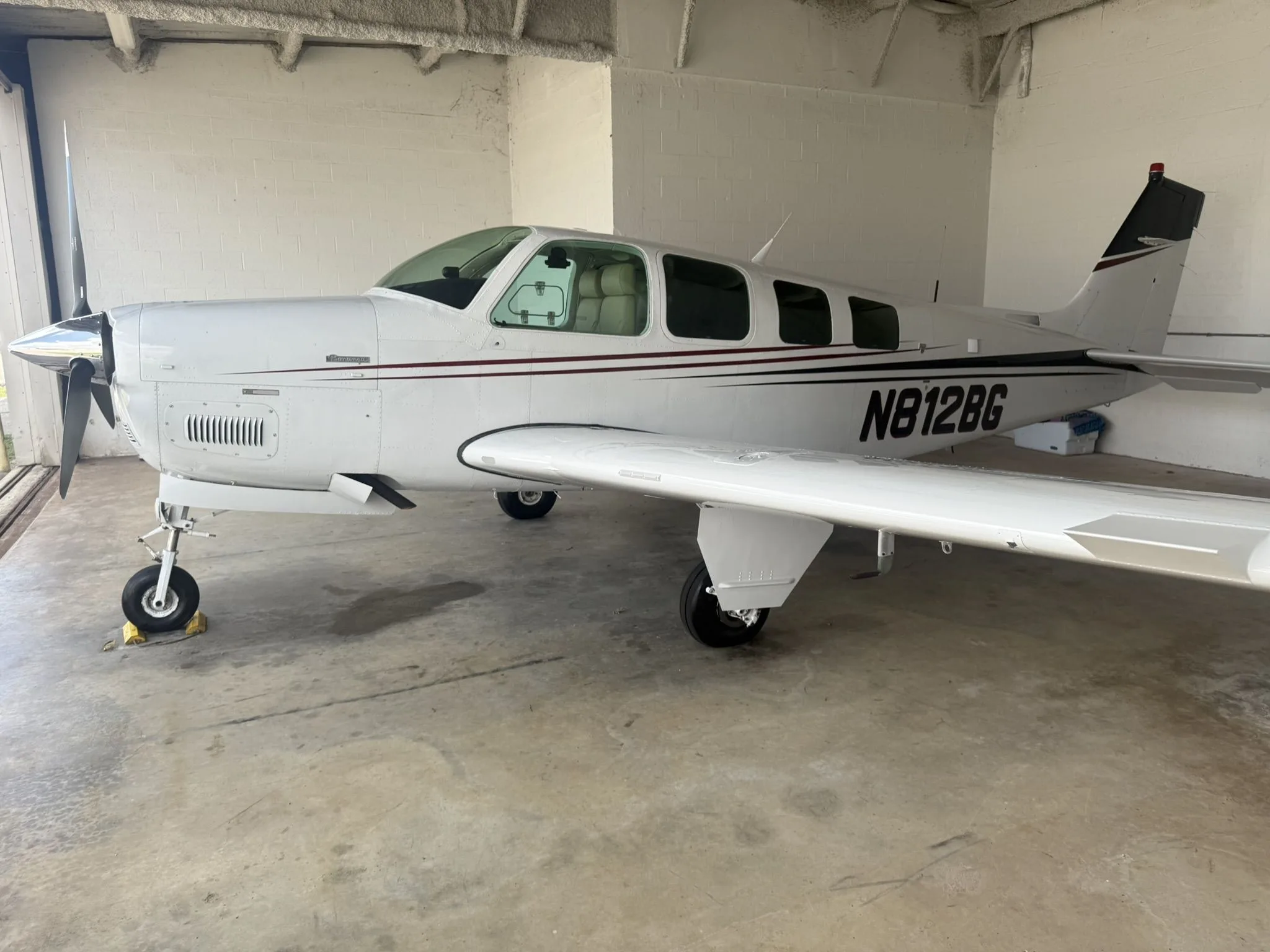 White Beechcraft Bonanza N812BG with dark red and black stripes parked inside a hangar.