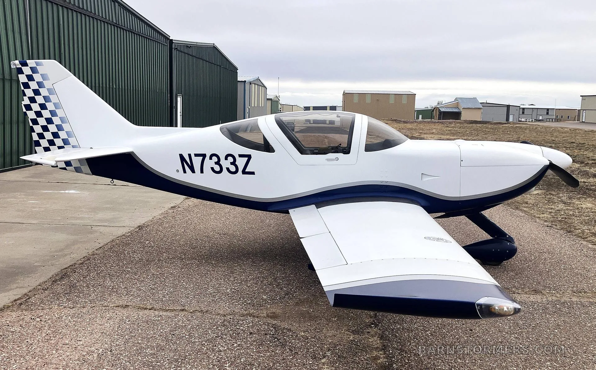 A white and blue single-engine aircraft with registration N733Z parked on an airfield next to hangars.