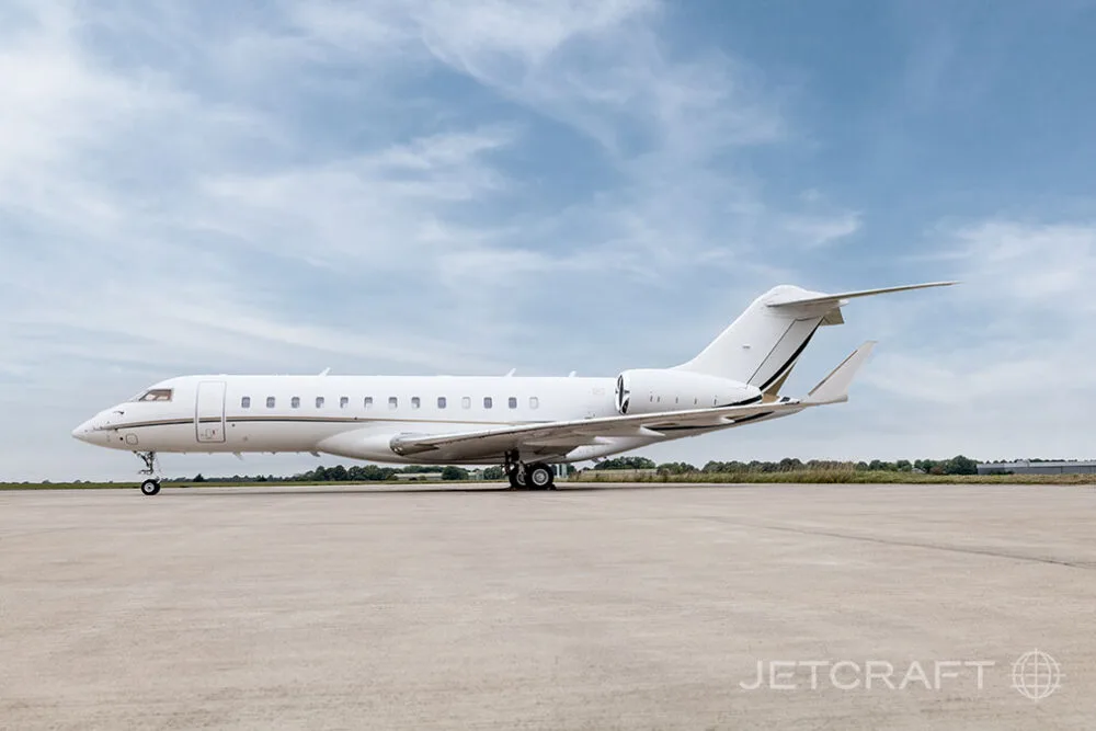 A white Bombardier Global business jet with gold and black stripes parked on an airfield, viewed from the side.