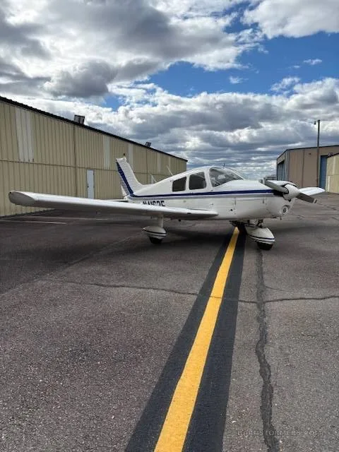 A white single-engine propeller aircraft with blue stripes (N4163F) parked on an asphalt tarmac next to hangars under a cloudy sky.