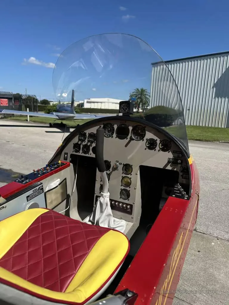 The cockpit of a red and yellow ultralight aircraft, showing the instrument panel, controls, and a red and yellow quilted seat.