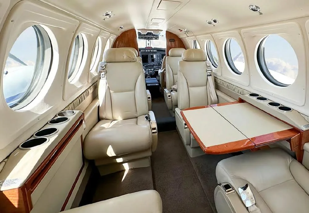The luxurious interior cabin of an aircraft with beige leather seats, a wooden table, and large windows.