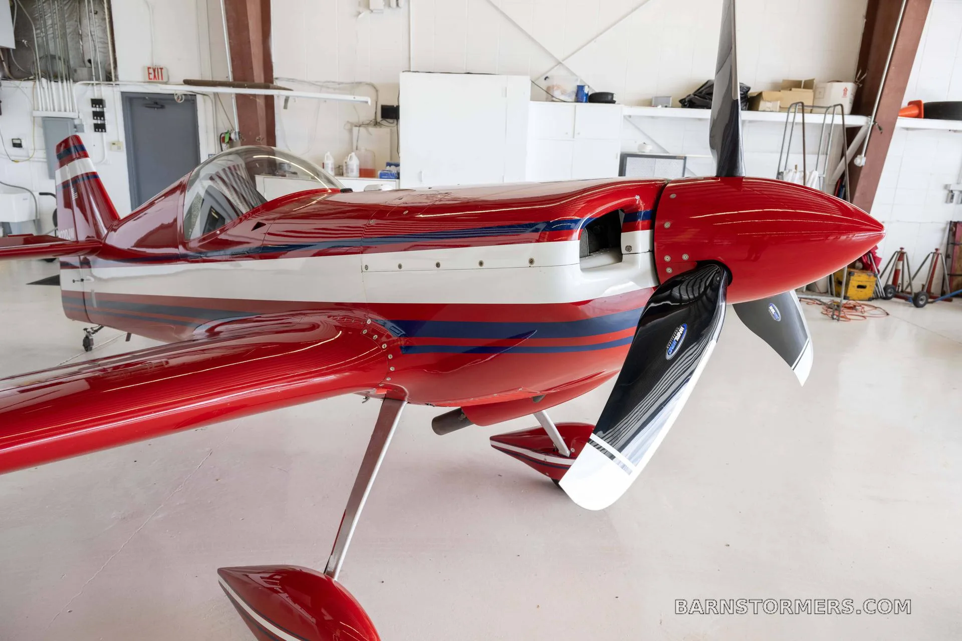 A close-up view of the nose and propeller of a red and white aerobatic aircraft in a hangar.