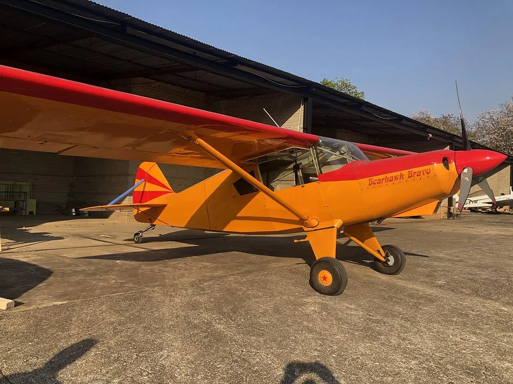 A red and orange Bearhawk Bravo aircraft is parked on a concrete pad in front of an open hangar on a sunny day.
