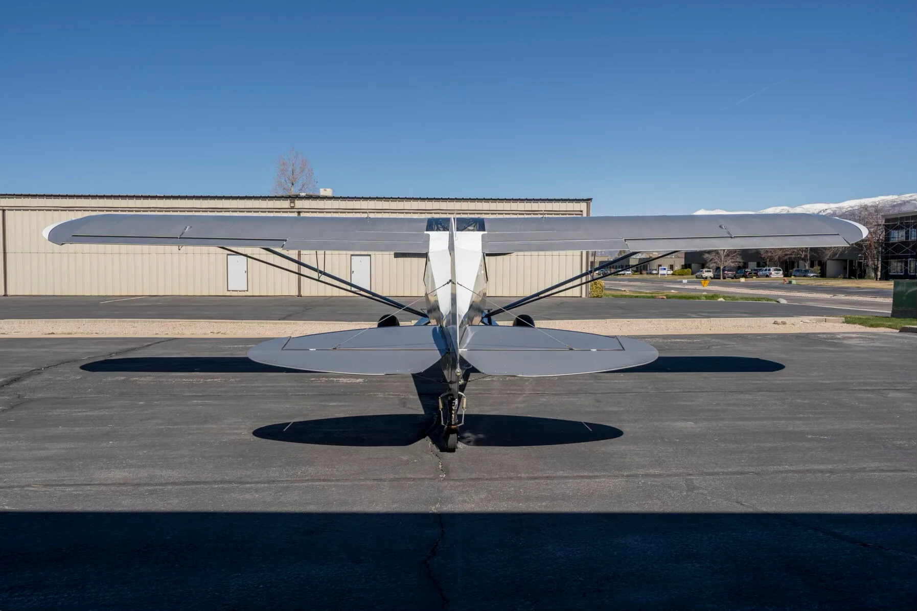 A rear view of the white and grey Carbon Cub FX-3 aircraft N25FX parked on an asphalt ramp in front of a hangar.