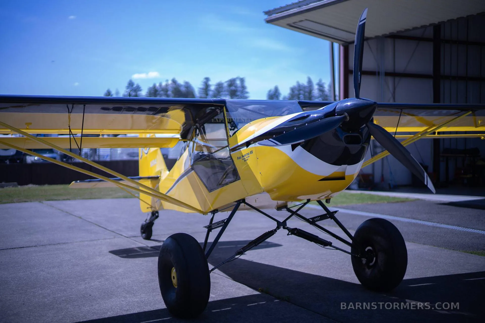 A yellow and black Kitfox aircraft with large tundra tires is parked on a tarmac in front of a hangar.