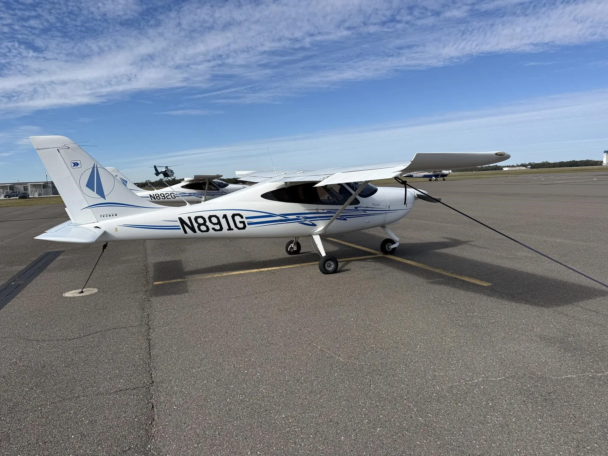 A white Tecnam P2008 aircraft with blue stripes and N891G registration, viewed from the front-right on an airport tarmac.