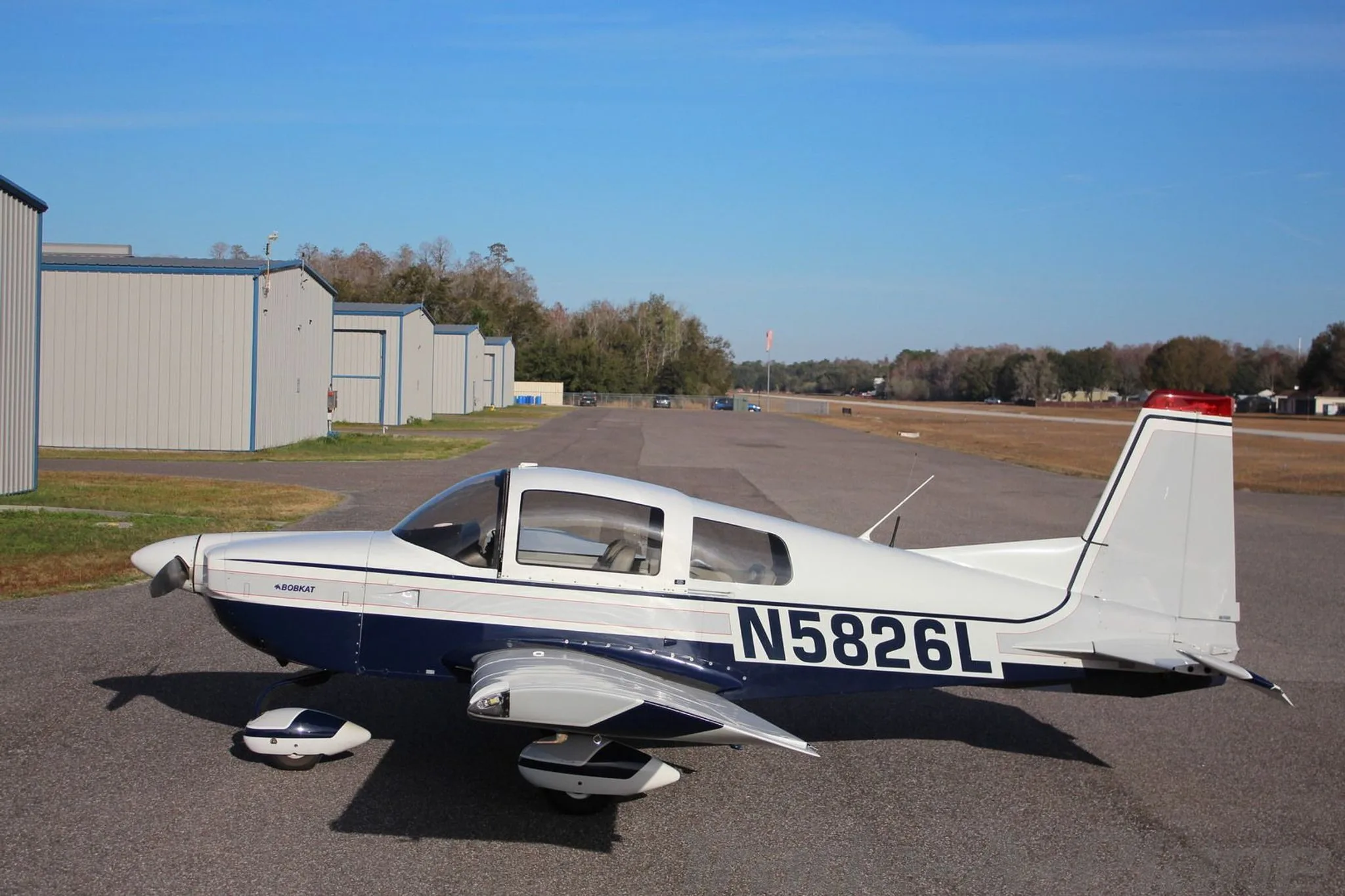 A white and blue Grumman AA-5B Tiger aircraft (N5826L) parked on an airfield with hangars in the background.