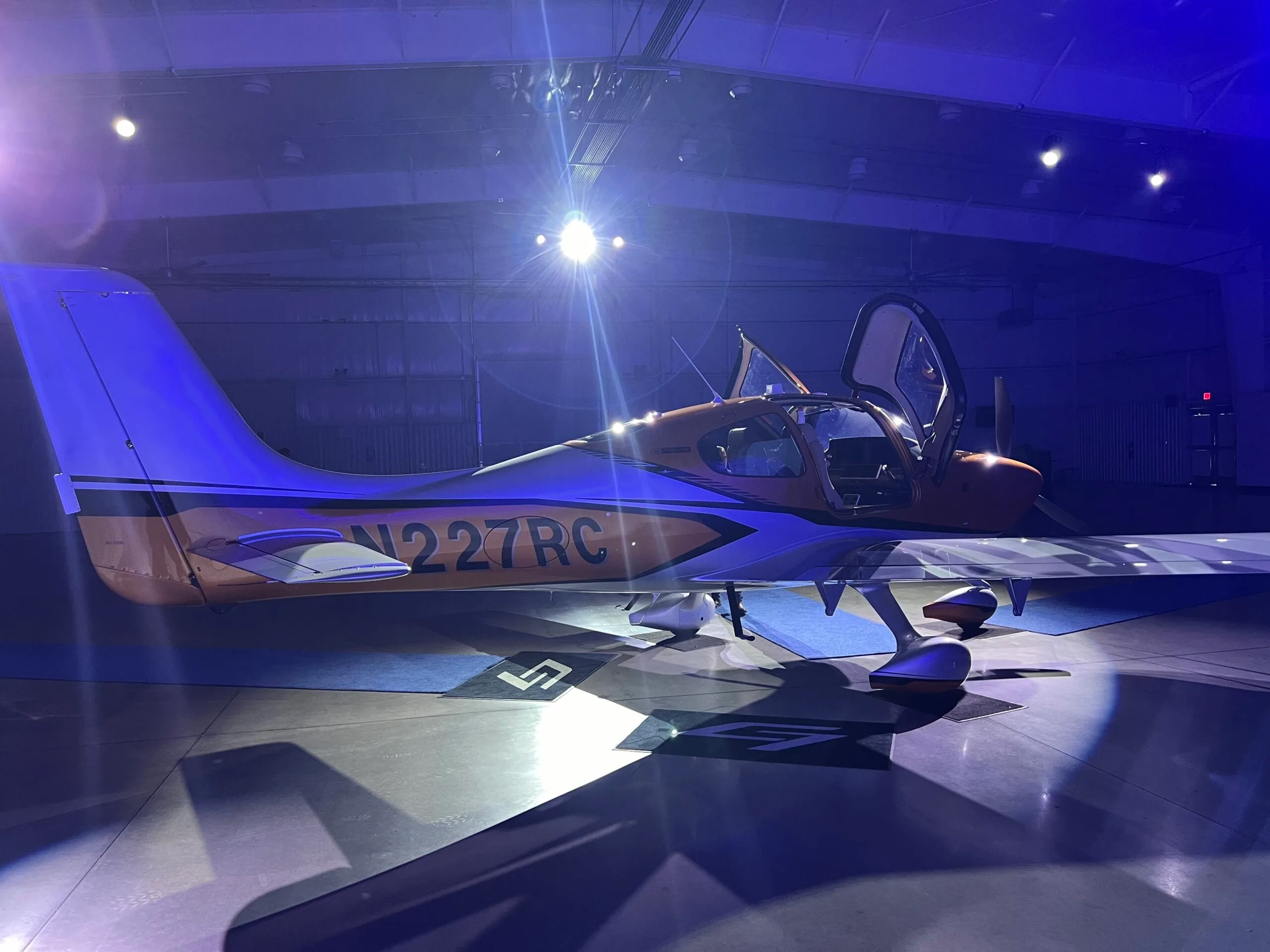 A side view of a Cirrus SR22 aircraft with orange and silver paint, doors open, illuminated by blue light in a hangar.