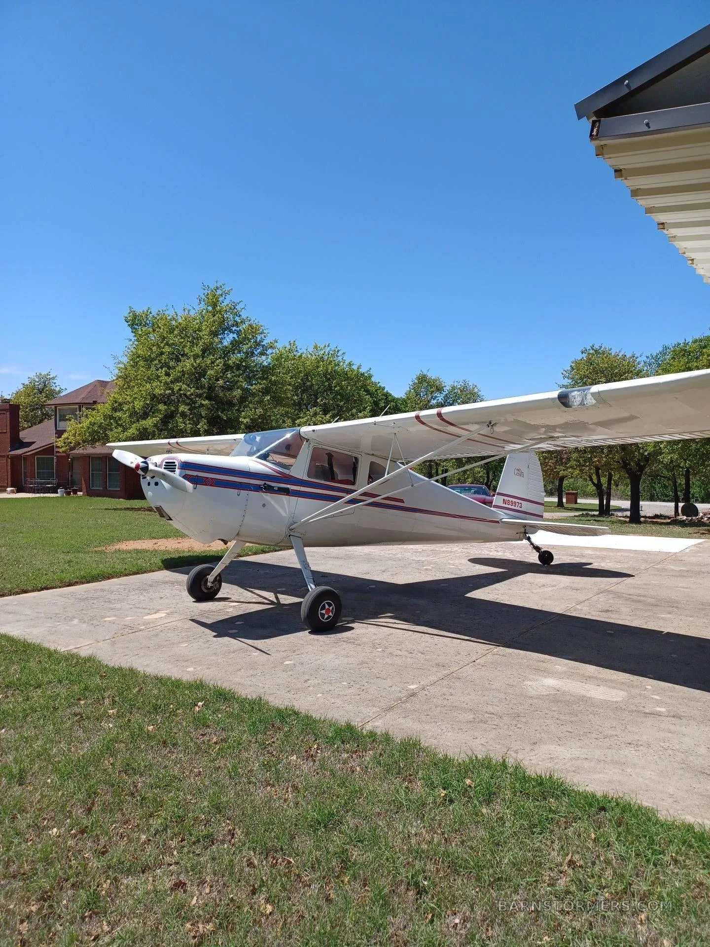 A white high-wing taildragger aircraft with blue and red stripes, tail number N6297D, parked on a concrete pad on a sunny day.