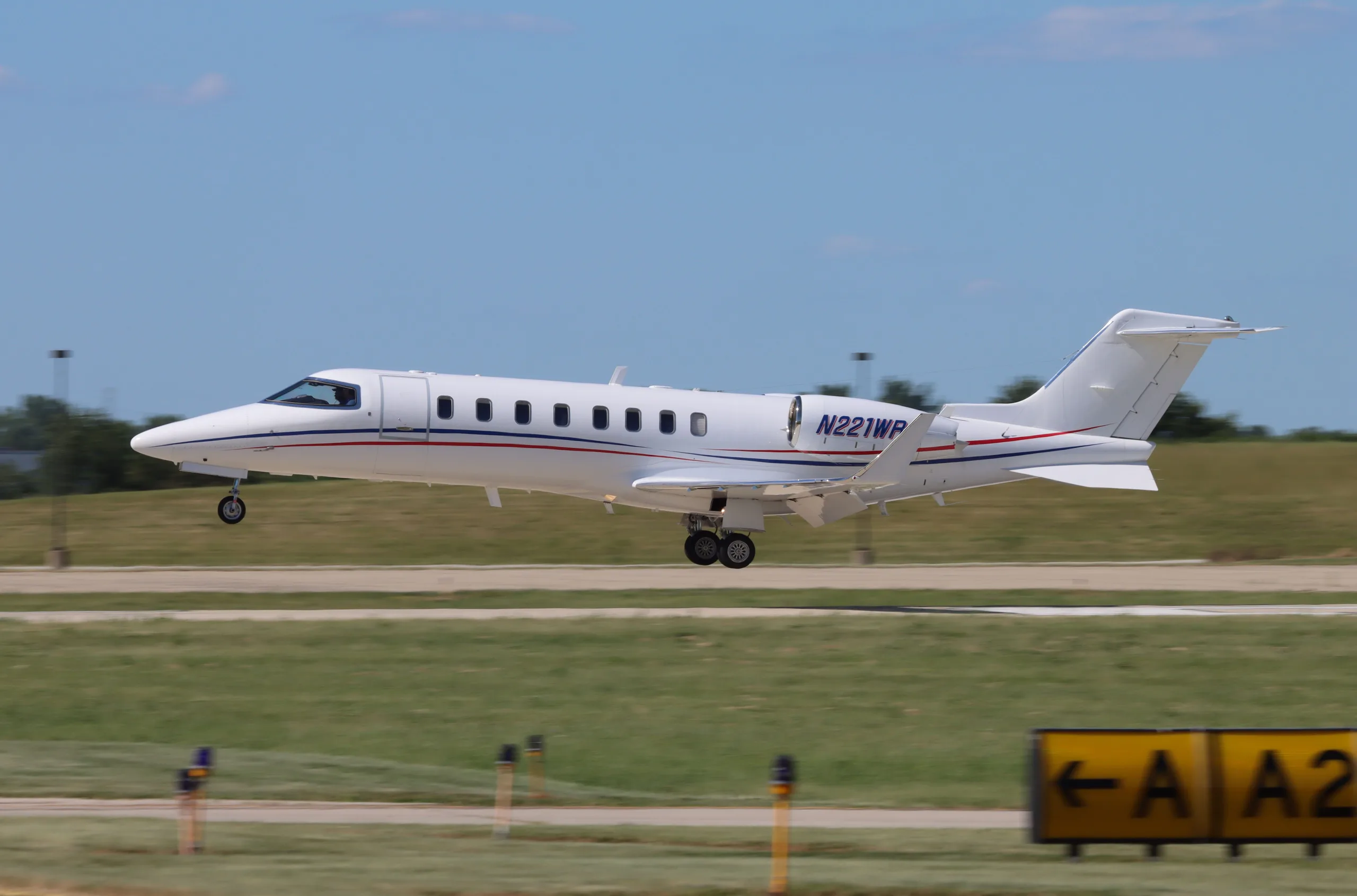 A white business jet with blue and red stripes, registration N221WP, is captured mid-air just above a runway with its landing gear extended.