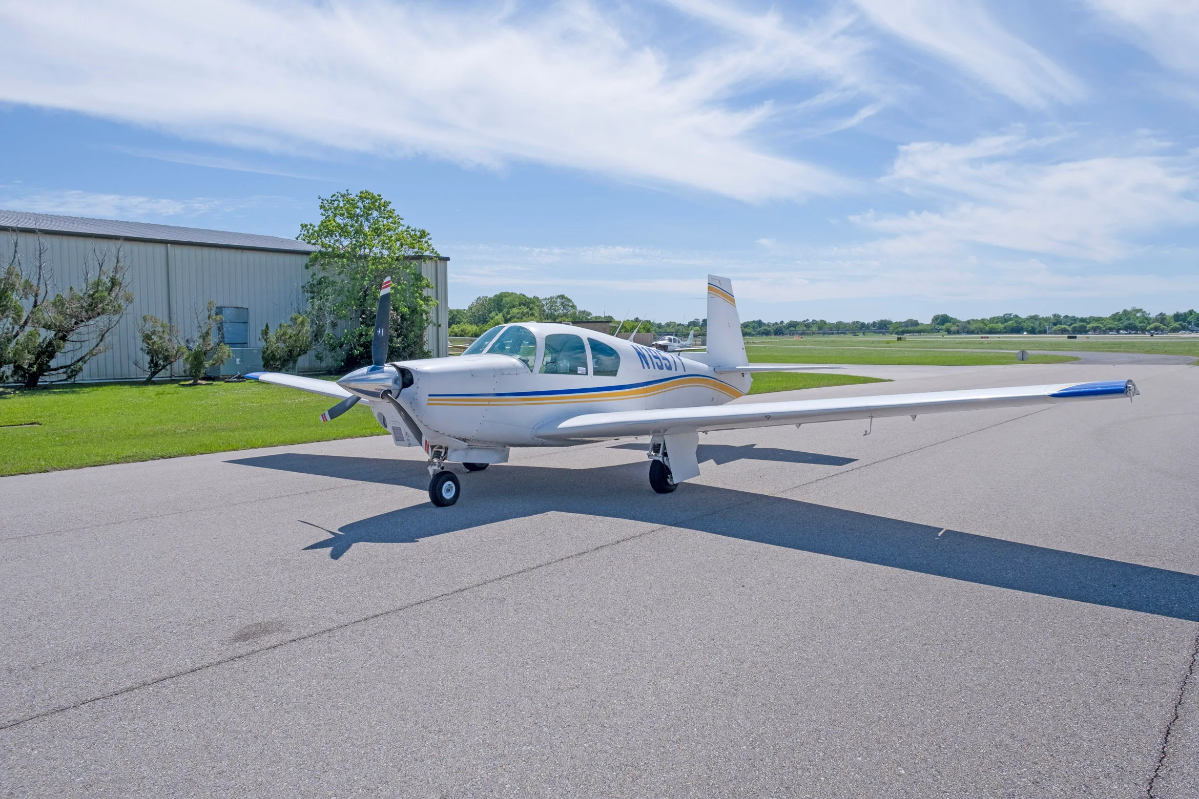 A white single-engine propeller aircraft with blue and yellow stripes, registration N1957Y, parked on an asphalt apron next to a hangar.