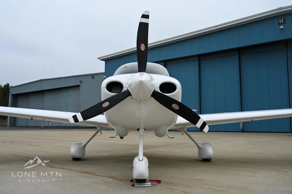 Front view of a white Cirrus SR22 aircraft with a three-bladed propeller, parked on an apron.