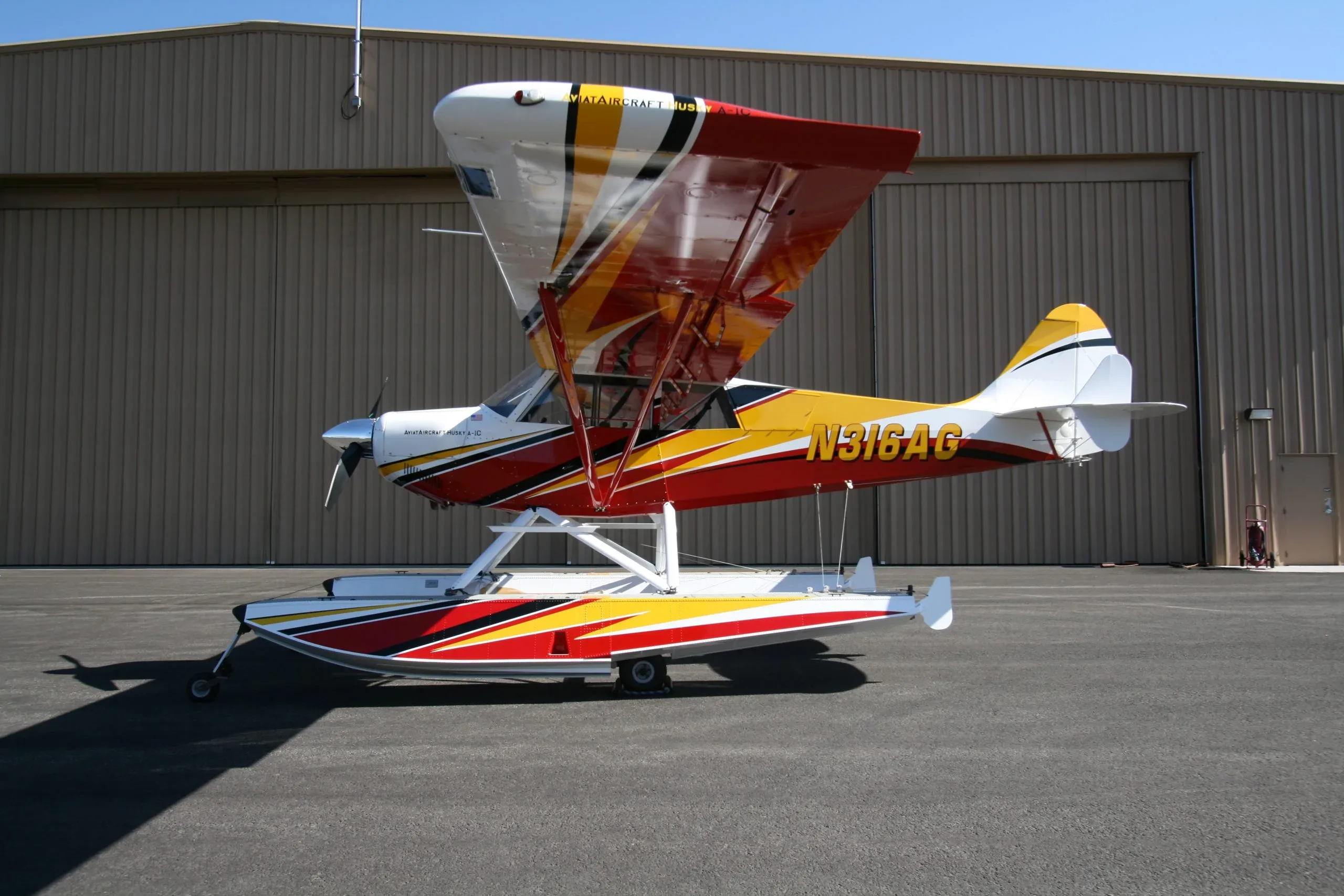 A red, yellow, and white Aviat Aircraft Husky A-1C seaplane, N316AG, parked on an asphalt ramp in front of a hangar.
