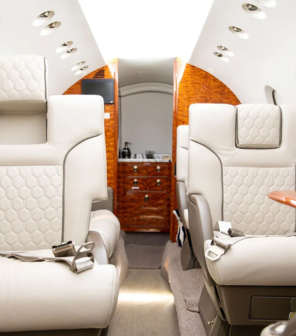 A view down the aisle of a private jet cabin, showing light grey quilted seats and a rear galley with polished wood cabinetry.