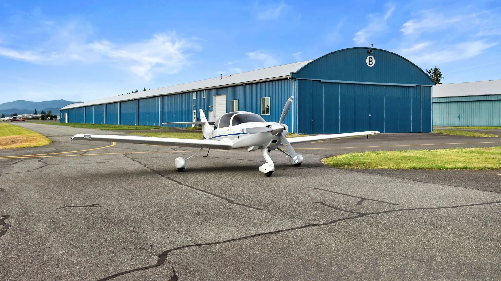 A white single-engine aircraft with blue stripes on the fuselage and tail, registration N404CB, parked on an asphalt runway with a long blue hangar in the background.