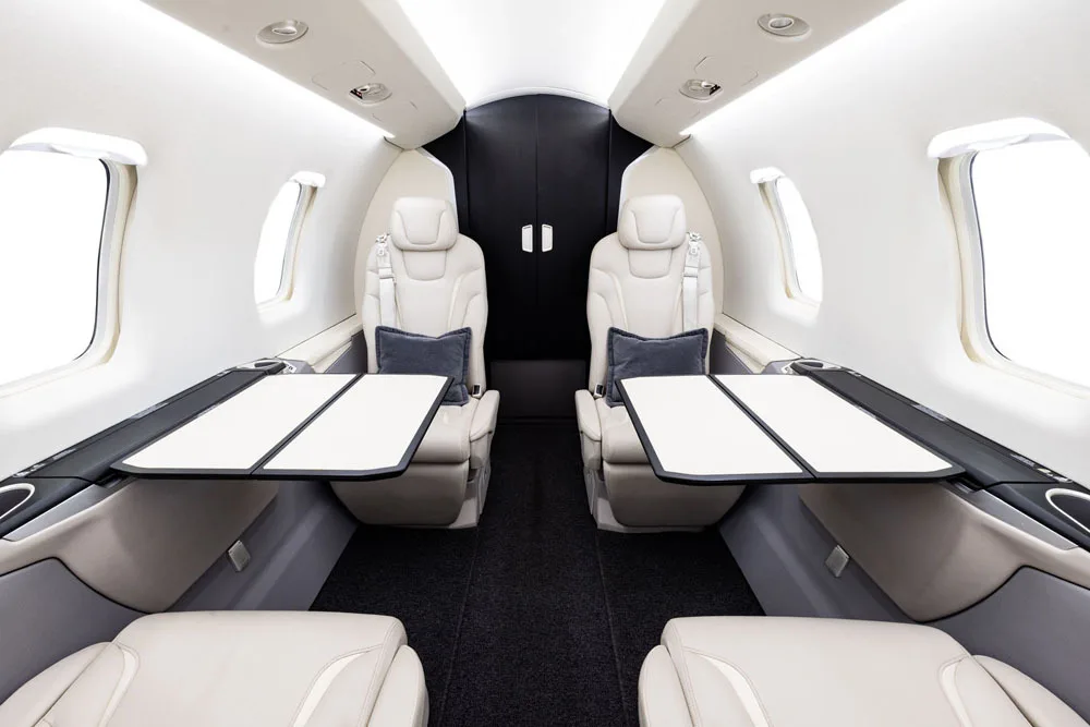 A private jet cabin interior showing two rows of light beige seats with tables extended and a dark partition at the rear.
