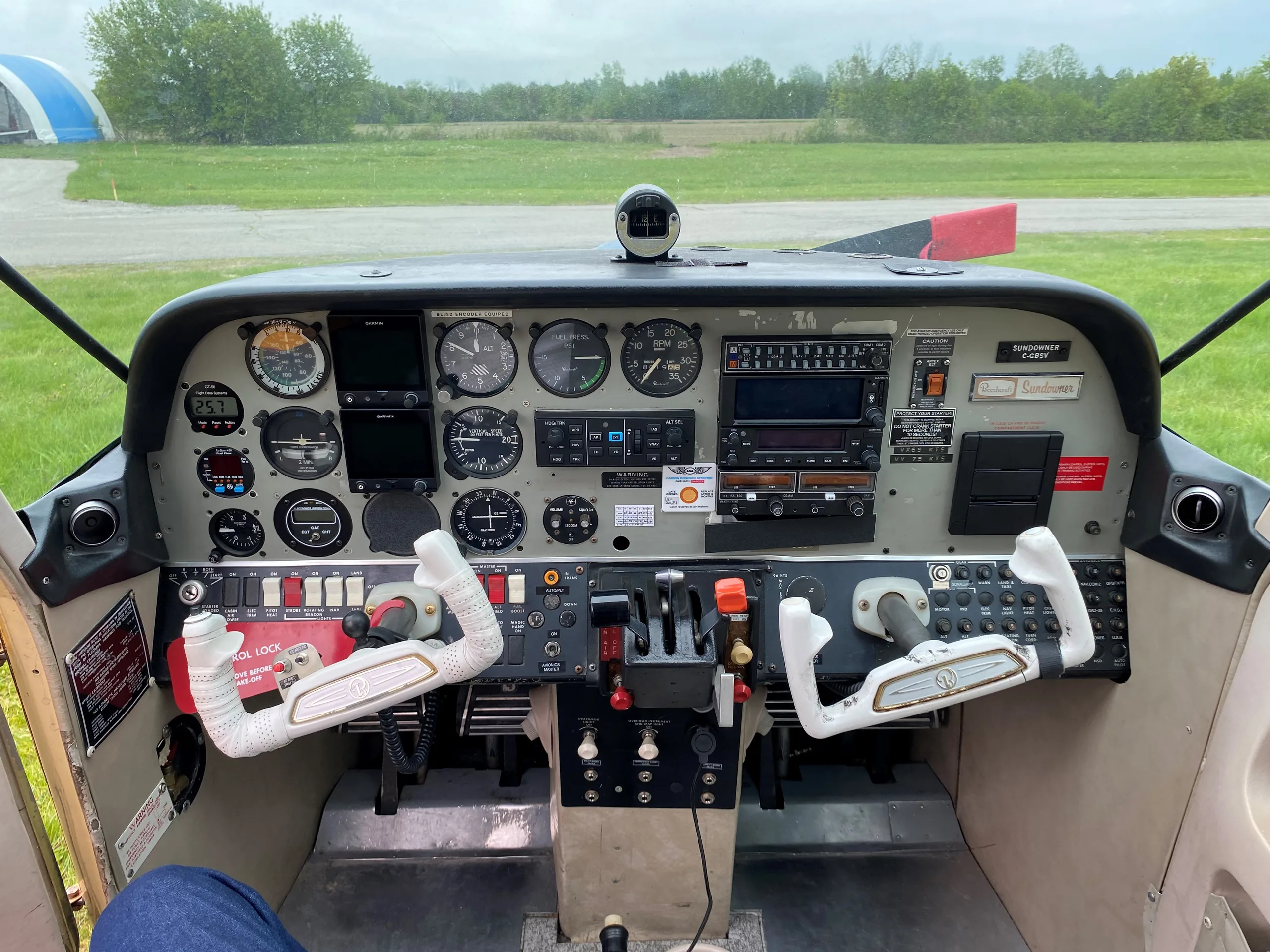 The cockpit of a Sundowner C-23 aircraft showing the instrument panel, controls, and a view of a grassy field outside.