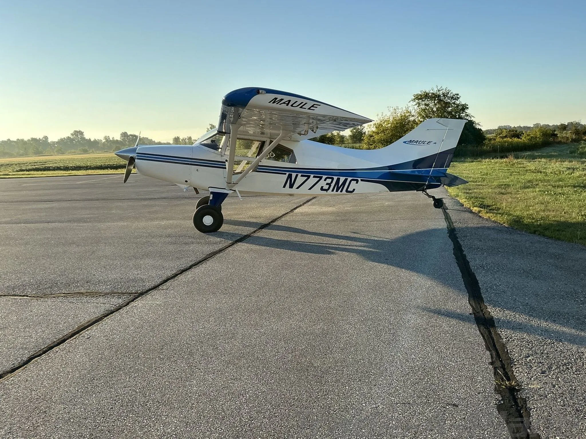 A side view of a white Maule aircraft with blue stripes, parked on an asphalt runway under a clear sky.