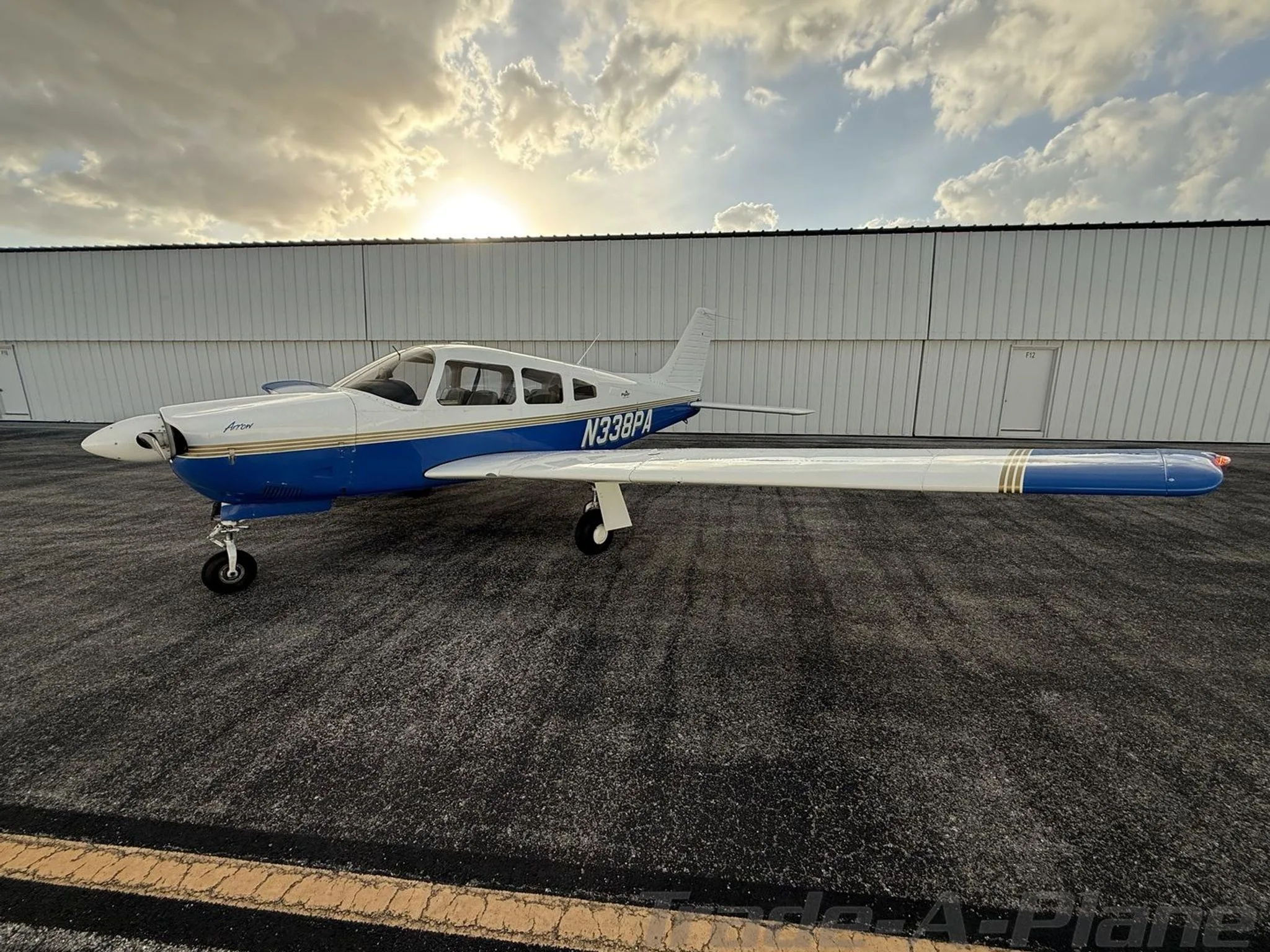 A white and blue Piper Arrow aircraft (N338PA) parked on the tarmac next to a hangar at sunset.