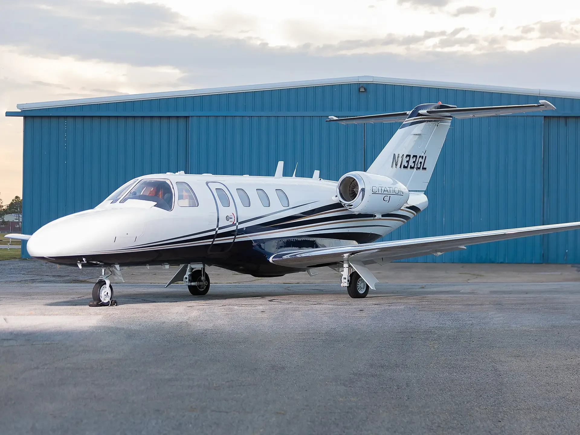 A white Cessna Citation CJ business jet with black and gold stripes is parked on an asphalt ramp in front of a blue hangar.