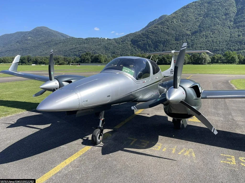 A silver Diamond DA42 aircraft with registration HB-LKN parked on an airfield, viewed from the front with mountains in the background.