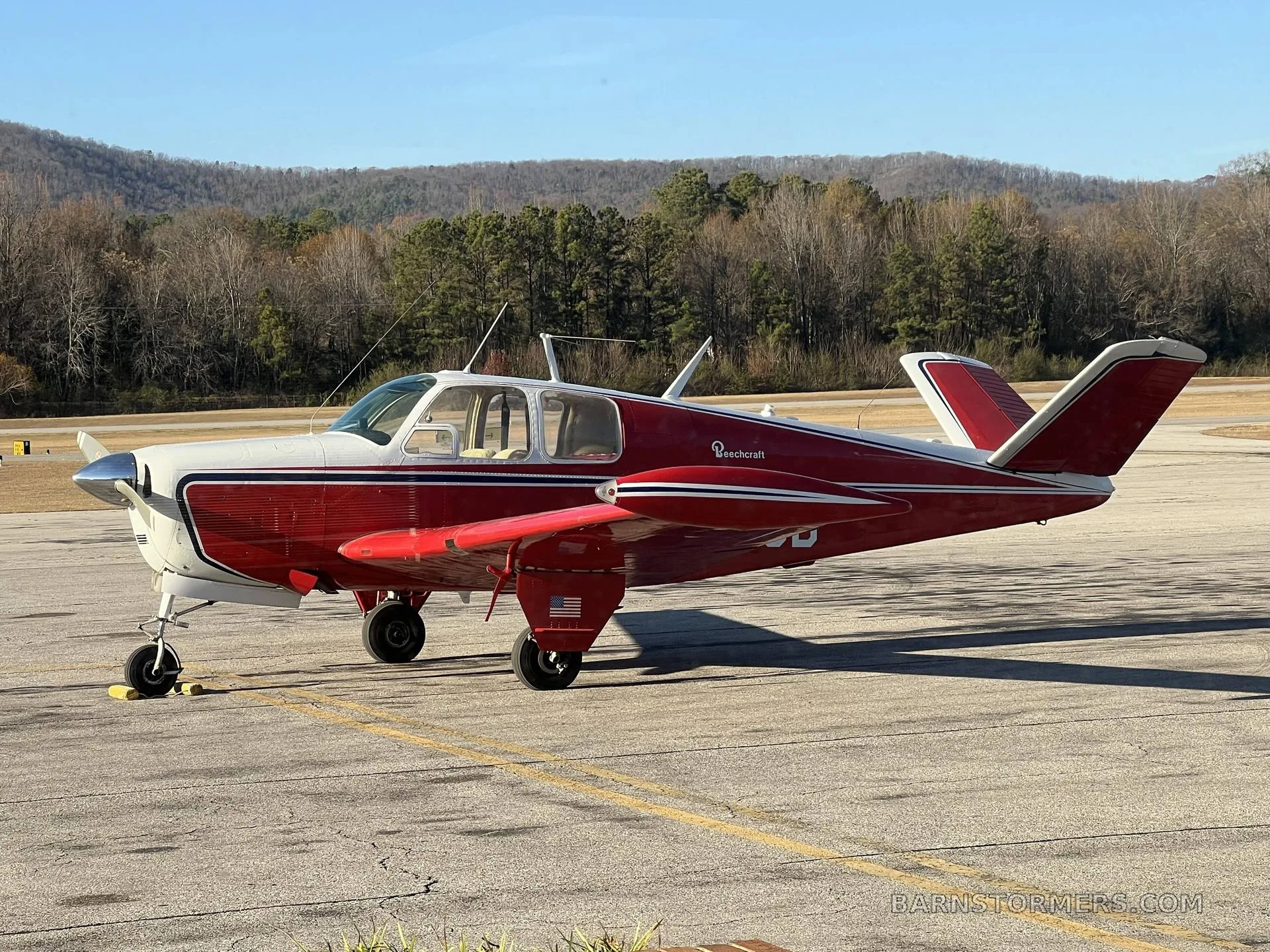 A red and white Beechcraft Bonanza is parked on an asphalt ramp with hills and trees in the background.