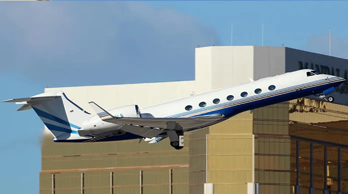 A white Gulfstream business jet with a blue stripe is seen taking off or landing against a backdrop of modern buildings.