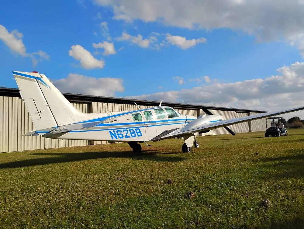 A white Beechcraft Baron N62BB with blue stripes is parked on a grassy field with a hangar in the background, viewed from the rear-left.