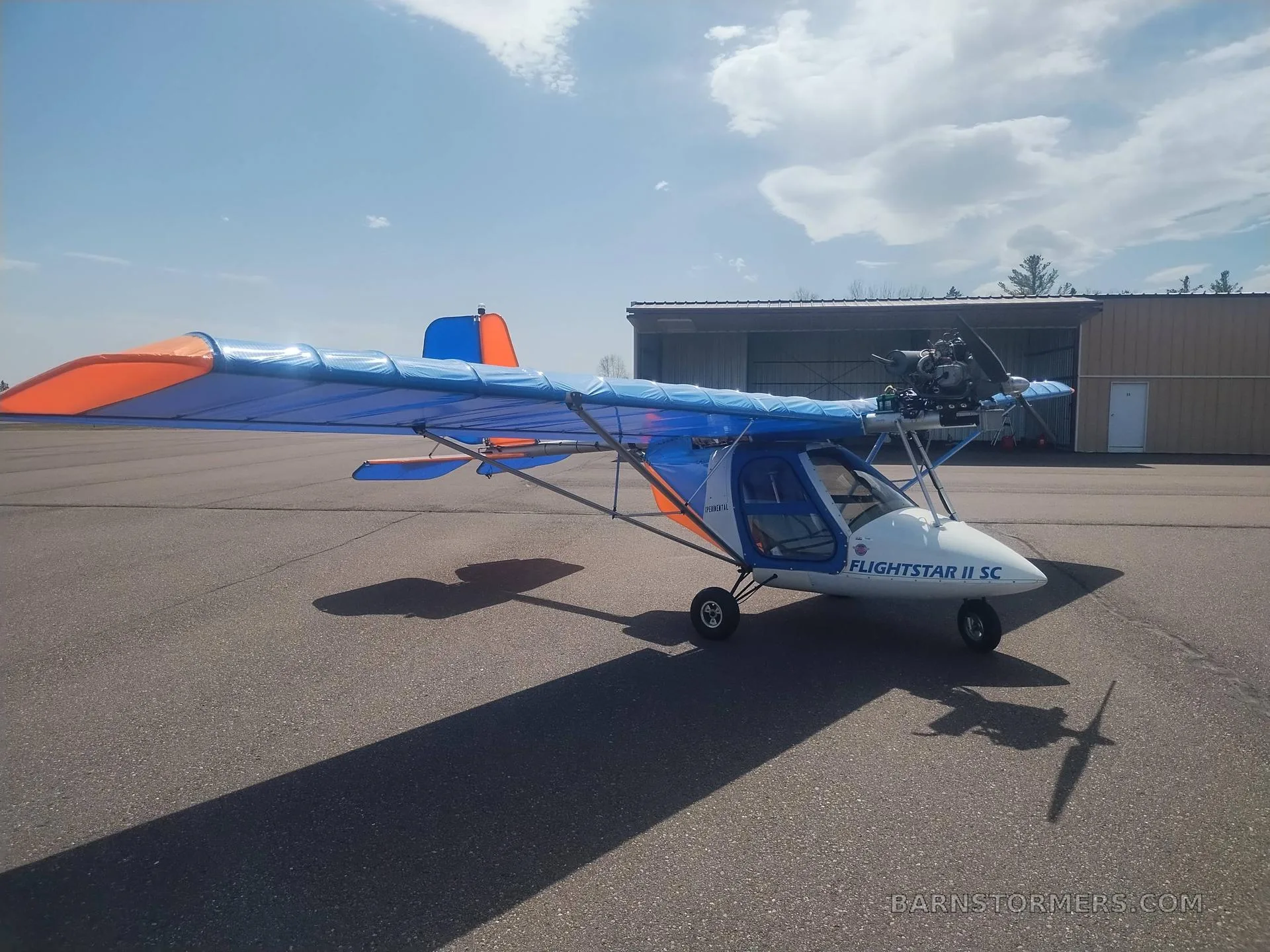 A blue and orange Flightstar II SC ultralight aircraft parked on an asphalt runway with a hangar in the background.