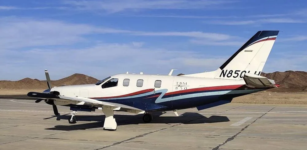 A white TBM 850 aircraft with blue and red stripes on the fuselage and tail, parked on an airfield with mountains in the background.