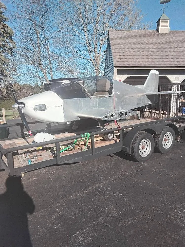A partially assembled, unpainted light aircraft with a white nose cone, secured on a black utility trailer in a driveway.