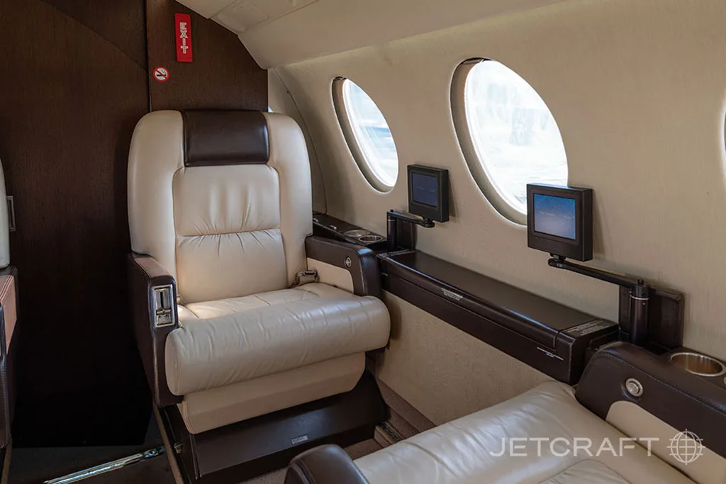 A close-up of a cream leather seat in the business jet cabin with a dark wood side console and entertainment screens.