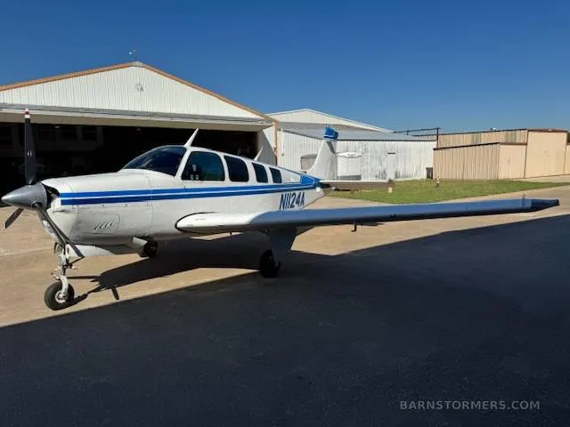 A white and blue Beechcraft Baron N1124A parked on an airfield with hangars in the background.