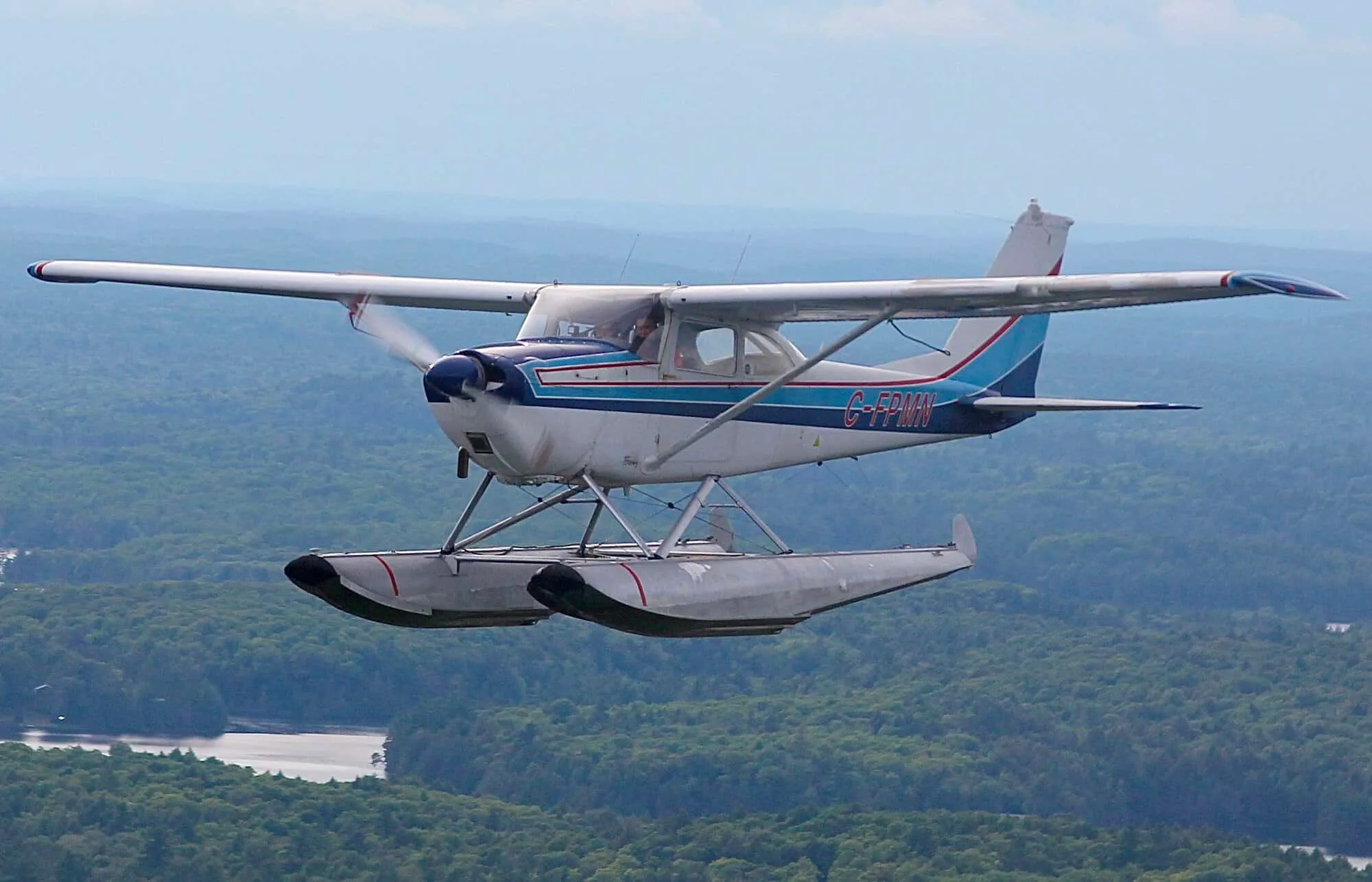 A white and blue Cessna 172 floatplane, registration C-FPMN, flies over a forested landscape with water below.