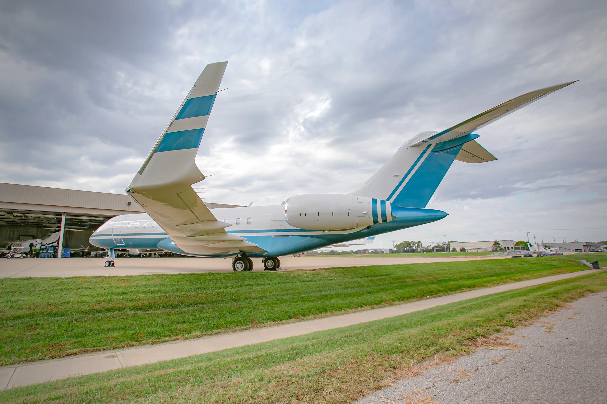 A rear view of a white and blue business jet, likely a Bombardier Global, with its tail visible, parked near a hangar.