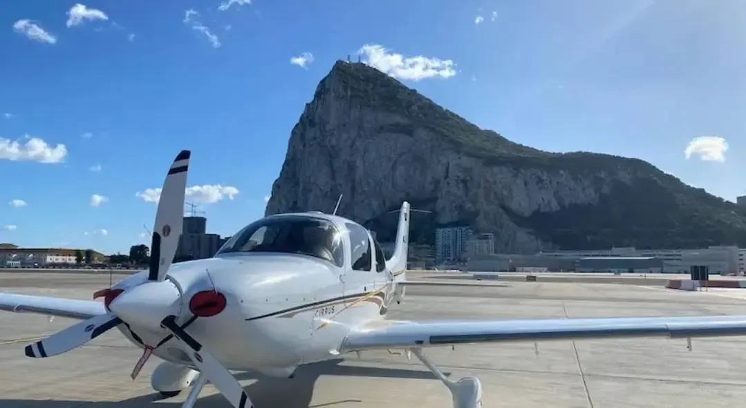A white Cirrus SR22 aircraft with gold and black stripes parked on an airfield with the Rock of Gibraltar in the background under a clear blue sky.