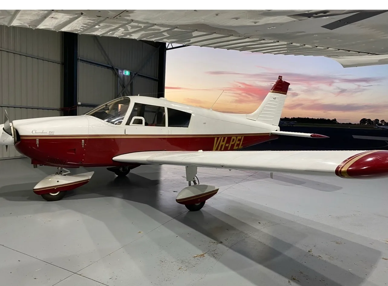 A red and white Piper Cherokee 180 aircraft, registration VH-PEL, is parked in a hangar with a sunset sky in the background.