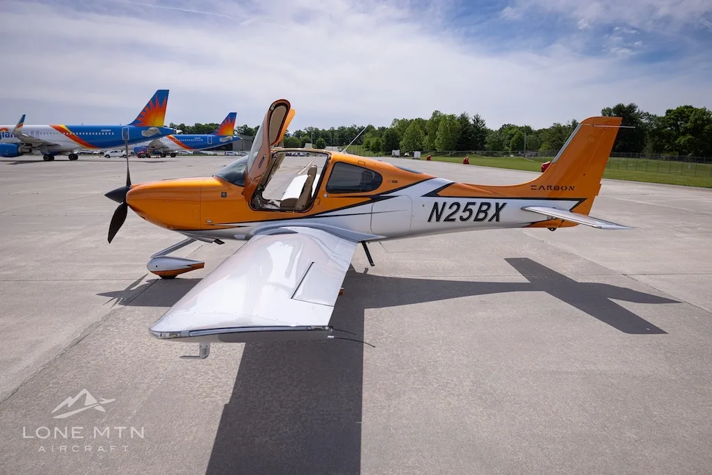 A rear-right view of the orange and silver Cirrus SR22T N25BX with its doors open on an airfield.